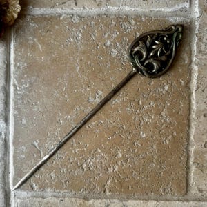 May include: A silver hair stick with a decorative, ornate, leaf-shaped top. The top is detailed with a swirling, floral design.