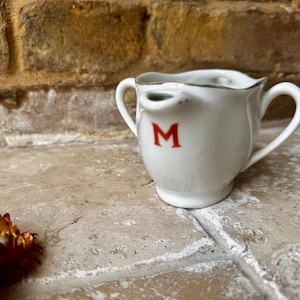 Puede incluir: Crema de cerámica blanca con una letra "M" roja en el lateral. La crema tiene un pico y dos asas.