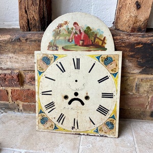 Puede incluir: Cara de reloj antiguo con números romanos y una escena pintada de una mujer en un jardín. La cara del reloj es blanca con números romanos negros y un borde amarillo. La escena está pintada en colores brillantes y representa a una mujer con un vestido rojo de pie en un jardín.