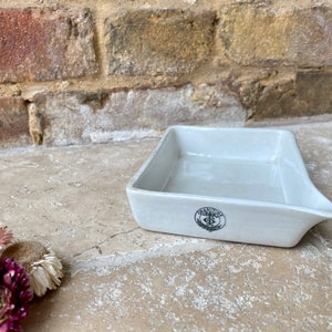 May include: A white ceramic rectangular dish with a small spout on one side. The dish has a black logo with the text "GRATTAGE" and a symbol.