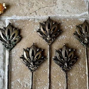 May include: Set of five gold-toned metal decorative floral-shaped finials with intricate details. Each finial has a long, thin stem.