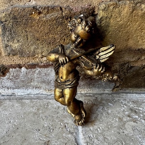 Puede incluir: Figura de querubín dorada tocando el violín. El querubín tiene el pelo rizado y alas, y está vestido con una prenda drapeada. La figura está montada en una pared de ladrillo texturizada. El querubín es un objeto decorativo.