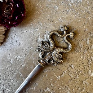 May include: A silver-toned letter opener with an ornate handle featuring a double serpent design. The handle has intricate scrollwork and floral accents. The opener is set against a textured, neutral-toned surface.
