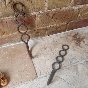 May include: Two dark brown, hand-forged metal hooks, each featuring a vertical design composed of three interconnected circular loops that taper into a single straight shank. The metal has a textured, weathered appearance with visible oxidation.