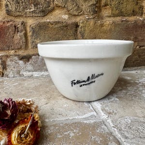 Puede incluir: Un cuenco de cerámica blanco con el texto "Fortnum & Mason at Piccadilly" en escritura negra. El cuenco está colocado contra una pared de ladrillo y una superficie de piedra. Flores secas en primer plano.