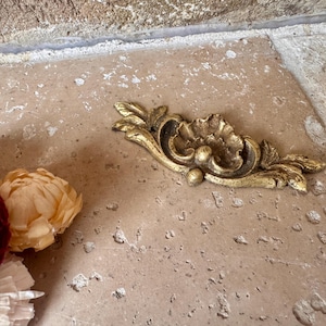 May include: A gold-colored decorative furniture applique featuring a central shell motif flanked by symmetrical leaf patterns. The piece has a textured, weathered finish with dark highlights in the recessed areas, showcasing an ornate, curved design.