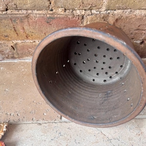 May include: A brown, cylindrical ceramic colander with numerous small holes in the bottom. The colander has a slightly flared rim and is set against a brick wall and stone surface. The color is a warm, earthy tone.