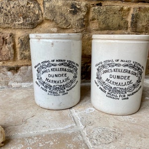 May include: Two white ceramic jars with black lettering. The jars are labeled "Grand Medal of Merit Vienna 1873", "James Keiller & Sons", "Dundee Marmalade", "Only Prize Medal for Marmalade", and "London 1883".