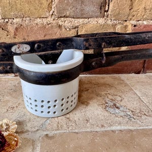 May include: Antique black potato ricer with a white ceramic bowl. The ricer has a lever-style handle and a perforated bowl for pressing potatoes. The metal shows signs of age and wear, adding to its vintage charm.
