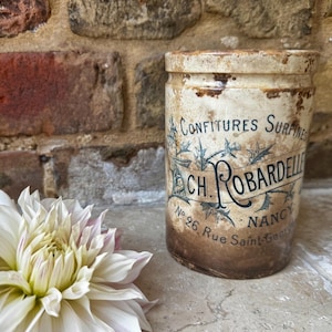 May include: A vintage cylindrical jar with distressed markings and text. The jar is labelled "CONFITURES SURFINES" and "CH. ROBARDELLE NANCY". A white and pink dahlia flower is in the foreground, with a brick wall in the background.