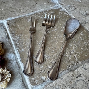 Puede incluir: Tres utensilios de servir de plata antiguos: dos tenedores y una cuchara. Los utensilios tienen mangos ornamentados y están dispuestos sobre una superficie de piedra. Hay flores secas en la esquina inferior izquierda. Los tenedores tienen tres y cuatro púas.