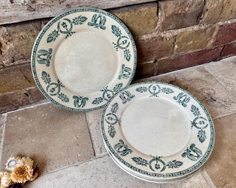 May include: Two antique white plates with a green floral and bird design around the rim. One plate is separate, and the other plates are stacked. The plates are on a stone surface with a brick background.