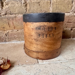 May include: A cylindrical wooden container with a natural light brown finish and a dark black painted rim. The side of the container features weathered black stencilled text that reads "DOUBT" and "LITRE". The base is reinforced with a wooden band.