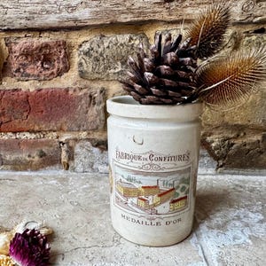 May include: A cream-coloured ceramic jar with the words "Fabrique de Confitures" and a factory illustration. The jar contains a pine cone and dried plants. The background is a brick wall. The jar is likely a vintage item.