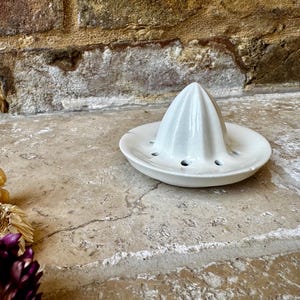 May include: White ceramic citrus juicer with a pointed top and a base with holes. The juicer is sitting on a light brown surface.