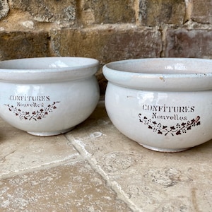 May include: Two white ceramic bowls with brown lettering that reads "Confitures Nouvelles". The bowls are sitting on a light brown tiled surface.