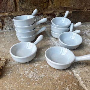 以下が含まれることがあります： 取っ手付きの小さな白いセラミック製サービングボウルのコレクション。ボウルは積み重ねられ、石の表面に配置されており、キッチンやダイニングの設定を示唆しています。ハンドルは湾曲しており、持ちやすくなっています。