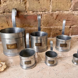 Op de afbeelding: Een set van zes trapsgewijze zilverkleurige metalen maatbekers met verticale handvatten. Elke beker is voorzien van een rechthoekig messingkleurig plaatje aan de voorzijde met het woord Lait in een gotisch lettertype in reliëf.