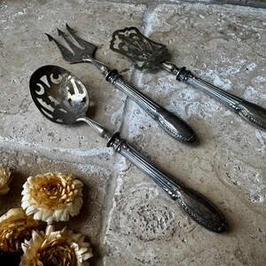 May include: Three antique silver serving utensils: a slotted spoon, a fork, and a server. The utensils have ornate handles and are arranged on a textured, light-coloured surface. Dried flowers are in the foreground.