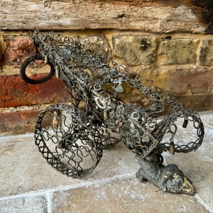May include: A decorative metal cannon made of wire and other metal pieces. The cannon has two wheels and a detailed design. The cannon is sitting on a stone surface.