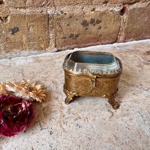 Può includere: Scatola portagioie antica dorata ornata con piano in vetro e piedini decorativi. La scatola rettangolare ha un coperchio incernierato e una chiusura. Una composizione di fiori secchi con fiori bordeaux e color crema è in primo piano.