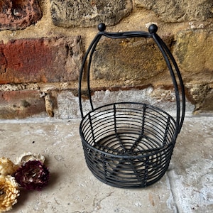 May include: A black wire basket with a handle. The round basket is made of vertical wires, with a handle arching over the top. The handle is attached with small ball-shaped details. Dried flowers in shades of cream and burgundy are in the foreground.