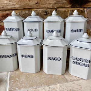May include: Set of eight white ceramic canisters with black trim and blue lettering. The canisters are labelled "raisins", "rice", "coffee", "salt", "currants", "peas", "sago", and "castor sugar".