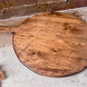 May include: A round, wooden cutting board with a handle. The board is made of light brown wood with visible grain and knots. The handle extends from the side. The board is approximately 16 inches in diameter.