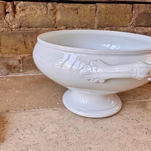 以下が含まれることがあります： 滑らかで光沢のある仕上げの白い陶器製スープチューリン。幅広で丸みを帯びたボウル、台座、両サイドに装飾的で彫刻が施されたハンドルが特徴です。この容器は、深い内部を備えたシンプルで洗練されたデザインとなっています。