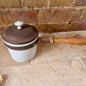 May include: A vintage white and brown ceramic saucepan with a lid and wooden handle. The lid is dark brown with a white knob. The handle is made of wood and metal. The saucepan is sitting on a stone surface.