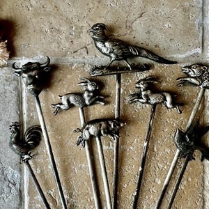 May include: A set of nine silver-toned metal skewers with animal figurines on top. The figurines include a rooster, a bull, a pig, two rabbits, a pheasant, a bird, and two wild boars.