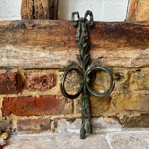 May include: An antique bronze-toned metal towel ring holder. The decorative piece features two circular rings, a bow, and leaf accents. The holder is mounted on a brick wall with a wooden beam.