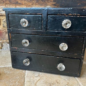 May include: A small, black wooden chest of drawers with four drawers. Each drawer has a clear, faceted glass knob. The chest has a distressed finish, with visible wear and tear. The background is a brick wall.
