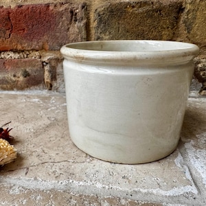 May include: A cream-coloured, cylindrical stoneware crock with a rounded rim. The antique container sits on a stone surface in front of a brick wall. Dried flowers are in the lower left corner.