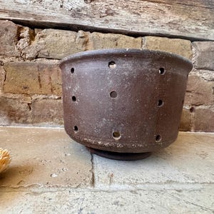 May include: A dark brown, cylindrical ceramic vessel featuring a series of evenly spaced circular perforations around its exterior walls. The bowl has a thick, rounded rim and a textured, matte finish, designed with a wide opening and a flat base.
