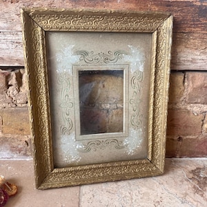 May include: A rectangular gold-toned picture frame featuring an ornate, embossed border with scrollwork patterns. The frame contains a light-colored mat with decorative, symmetrical line art and subtle white distressed accents surrounding a central opening.