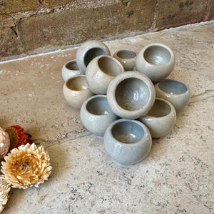 May include: A collection of small, round, light gray ceramic pots with a smooth, glossy finish. The pots have a circular opening at the top and are arranged in a cluster. Dried flowers in shades of red, orange, and cream are in the foreground.