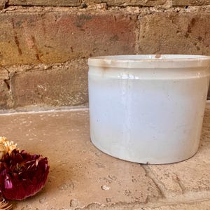 May include: A cylindrical ceramic crock with a smooth, off-white glazed finish. The vessel features a simple, straight-sided design with a slightly thickened rim at the top and subtle signs of wear, including minor discolouration near the upper edge.