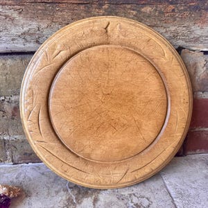 Peut inclure: Une assiette ronde en bois brun clair avec un motif de feuilles sculptées sur le bord. L'assiette a un centre surélevé et un grain de bois visible. Pour la décoration ou le service.