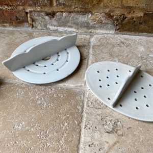 May include: Two light grey ceramic soap dishes. One is round with a raised, curved handle and concentric circles. The other is oval with a raised handle and multiple drainage holes. Both are on a stone surface.