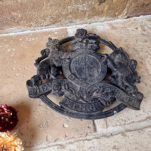 Op de afbeelding: Een ronde onderzetter van zwart gietijzer met het koninklijk wapen van het Verenigd Koninkrijk. Het ontwerp toont een leeuw en een eenhoorn die een centraal schild flankeren, bekroond met een kroon en een banier met de tekst DIEU ET MON DROIT.