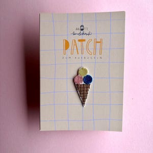 Könnte beinhalten: Ein gestickter Aufbügler in Form einer Eiswaffel mit drei Eiskugeln in Gelb, Rosa und Blau, befestigt auf einer beigen Karte mit blauem Gittermuster. Die Karte trägt den Text "PATCH ZUM AUFBÜGELN" sowie ein kleines Logo im oberen Bereich.