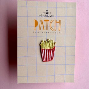 Könnte beinhalten: Ein gestickter Aufbügler in Form einer Pommes-Frites-Tüte mit goldgelben Pommes und einem rot-weiß gestreiften Behälter. Der Aufnäher ist auf einer beigen, rechteckigen Karte mit blauem Gittermuster und dem Text PATCH ZUM AUFBÜGELN befestigt.