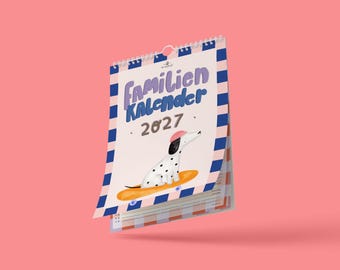 May include: A 2027 wall calendar with a spiral binding, featuring a white dalmatian dog wearing a pink hat on an orange surfboard. The cover has a blue and pink striped border and the text "Familien Kalender 2027" in purple and blue lettering.