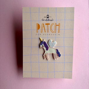 Könnte beinhalten: Ein gestickter Einhorn-Aufnäher mit gelbem Horn, violetter Mähne und weißen Flügeln auf einer beigen Karte mit blauem Gittermuster. Die Karte trägt die Aufschrift "kindsköpfe", "PATCH" und "ZUM AUFBÜGELN" in schwarzer und oranger Schrift.