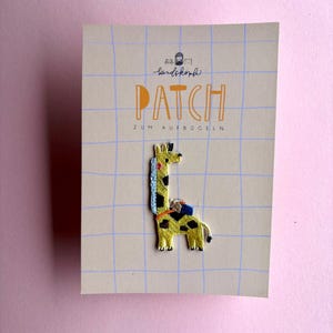 Könnte beinhalten: Ein gestickter Giraffen-Aufnäher mit gelbem Fell, schwarzen Flecken, blauer Mähne und einem kleinen blauen Rucksack, befestigt auf einer beigen Karte mit blauem Gittermuster, dem Text "PATCH ZUM AUFBÜGELN" und einem kleinen Logo oben.