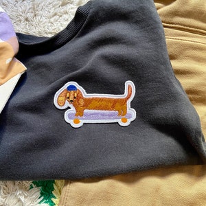 Könnte beinhalten: Ein dunkelgraues Sweatshirt mit Rundhalsausschnitt und einem gestickten Aufnäher eines braunen Dackels, der eine blaue Mütze trägt und auf einem hellvioletten Skateboard mit orangefarbenen Rollen steht. Das Sweatshirt liegt neben einer beigen Hose.