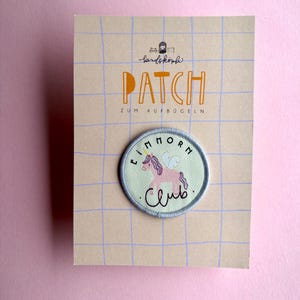 Könnte beinhalten: Ein runder Aufbügel-Patch mit einem rosa Einhorn, weißen Flügeln und einem gelben Horn auf hellgrünem Grund. Der Schriftzug "EINHORN CLUB" ist um das Einhorn gestickt. Der Patch ist auf einer beigen Karte mit blauem Gittermuster befestigt.