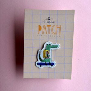 Könnte beinhalten: Ein gestickter Aufbügler in Form eines grünen Krokodils mit gelbem Rucksack und gestreiftem Shirt auf einem blauen Skateboard. Der Aufnäher ist auf einer beigen, rechteckigen Karte mit blauem Gittermuster und dem Text PATCH ZUM AUFBÜGELN befestigt.