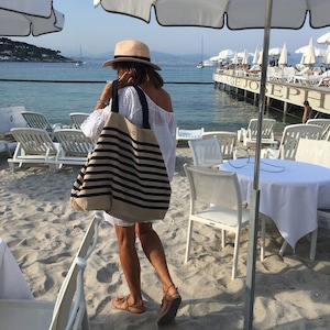 May include: A woman in a white crochet dress and a straw hat carries a striped tote bag on a beach. The bag has a black and white striped pattern and the text "LG" is visible on the front.